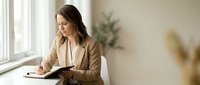 Professional businesswoman in beige blazer writing notes by window in modern office space for corporate planning and productivity concepts.