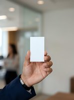 Male professional holding blank white business card in modern office environment with blurred colleagues and glass partitions in background.