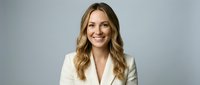 Professional blonde businesswoman in white blazer smiling confidently against gray background for corporate headshot and marketing materials.
