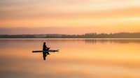Lone kayaker paddling on calm lake at golden sunrise with perfect reflection, peaceful morning water sport adventure creating serene outdoor recreation moment.