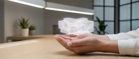 Young woman with light skin tone holding glowing holographic cube above wooden desk in modern office environment with plants and windows.