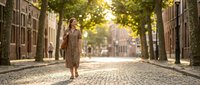 Young Caucasian woman in linen dress walking on sunlit cobblestone street lined with trees, carrying leather bag, smiling confidently.