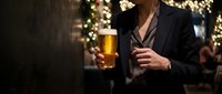 Man in dark business suit holding glass of beer at festive holiday party with warm golden lights and decorated background, suitable for seasonal marketing, hospitality, celebration, and lifestyle content.