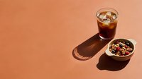 Iced cola beverage with ice cubes next to bowl of granola cereal on warm peachy background with natural shadows, suitable for food and beverage advertising.