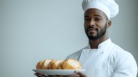 Young African American male chef in white uniform and hat holding freshly baked bread rolls on plate against neutral background, professional culinary portrait.