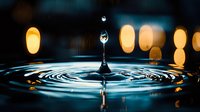 Water drop splashing on dark blue surface creating concentric ripples with golden bokeh lights in background, dramatic macro photography for wellness concepts.