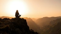 Woman meditating on mountain cliff at sunrise with golden light over canyon landscape for wellness and mindfulness concepts.