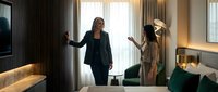 Professional businesswoman presenting luxury hotel suite to elegant female guest in upscale accommodation with modern interior design and premium amenities.