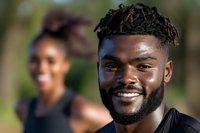 Young African American man with dreadlocks smiling outdoors with blurred woman in background during sunny day for lifestyle and diversity concepts.