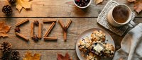 Cozy autumn flat lay with cinnamon sticks spelling cozy, oatmeal cookies, hot tea, pine cones, and fall leaves on rustic wooden background.