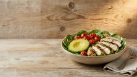 Grilled chicken breast slices arranged with fresh greens, tomatoes and avocado in a ceramic bowl on rustic wood surface for healthy meal prep and nutrition content.