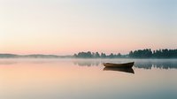 Solitary wooden boat floating on calm misty lake at sunrise, with forested shoreline reflected in still water. Serene landscape for nature, travel, and meditation projects.