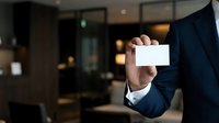 Businessman in dark suit holding blank white business card in modern office environment with blurred interior background for corporate branding and networking purposes.