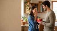 Young man surprising girlfriend with red gift box in cozy kitchen, woman covering mouth in excitement, romantic couple celebrating special occasion at home.