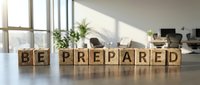 Wooden blocks spelling "BE PREPARED" on office desk with plants and windows in background, representing business readiness and planning for corporate environments.