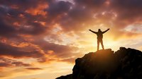 Person standing on mountain peak with arms raised celebrating success against dramatic orange sunset sky with clouds for motivational and achievement concepts.