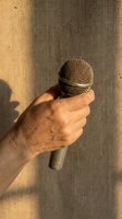 Hand holding microphone against textured wall with warm golden sunlight creating dramatic shadows for broadcasting and communication concepts.