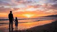 Adult male and young child standing together on sandy beach watching golden sunset over ocean waves, silhouetted against colorful sky for family bonding and travel concepts.