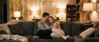 Young couple embracing on cozy sofa in warm living room with fairy lights creating romantic atmosphere for home lifestyle content.