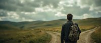 Man with backpack standing on rural dirt road facing mountains and rolling hills landscape under cloudy sky, outdoor adventure and hiking.