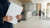 Businessman holding blank white document in modern office lobby with blurred colleagues and glass partitions in background for corporate communication.