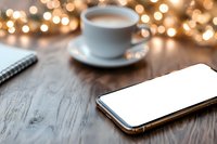 Smartphone with blank white screen on wooden table next to coffee cup and notebook, with soft bokeh lights creating cozy atmosphere. Mockup for app design.