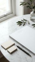 Minimalist workspace with marble surface, blank notebook, sticky notes, pen, and fresh olive branches in glass vase by window for creative projects and planning.