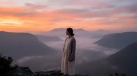 Woman in casual cardigan standing on mountain overlook at sunrise, gazing at misty valley below with layered mountain peaks in soft golden and pink light.