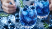 Refreshing blue cocktail with blueberries, ice cubes and rosemary garnish in glass. Sparkling summer drink with berries on dark background. Creative beverage photography.