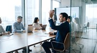 Professional businessman raising hand during corporate meeting in modern glass conference room with diverse team collaborating on business strategy.