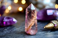 Pink quartz crystal point with natural inclusions on dark wooden surface, surrounded by purple stones and golden decorative elements with bokeh lights.