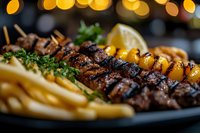 Grilled beef kebab with french fries and roasted yellow peppers on dark plate, garnished with fresh herbs and lemon slice. Bokeh lights background creates cozy atmosphere.