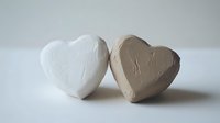 Two heart-shaped clay or plaster objects, one white and one beige, placed together against neutral background, showing texture and minimalist composition.