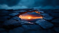 Dramatic sunset reflection captured in water puddle through broken ice surface, creating natural frame with dark blue winter atmosphere and orange glow.