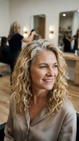 Mature blonde woman with curly hair smiling in modern hair salon during professional styling appointment with hairdresser working in background.