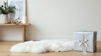 White fluffy faux fur rug on wooden floor next to wrapped gift box and wooden shelf with dried flowers and candles for home decor.