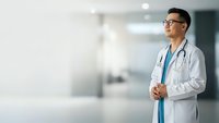 Asian male doctor in white coat with stethoscope standing confidently in modern hospital corridor, professional healthcare worker ready for patient care.