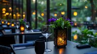 Elegant dining table setup with purple flowers in illuminated lantern, empty wine glass, and blurred restaurant interior with warm ambient lighting and garden views through windows.