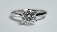 Classic solitaire engagement ring with round brilliant diamond in six-prong setting on polished platinum band against neutral gray background.