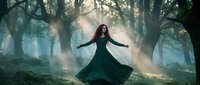 Mystical woman in flowing emerald dress dancing in enchanted forest with dramatic sunbeams filtering through ancient trees creating magical atmosphere.