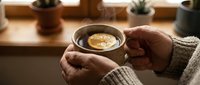 Person holding warm coffee cup with latte art in cozy home setting with potted plants on wooden shelf, perfect for lifestyle and beverage marketing.