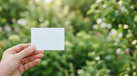 Hand holding blank white card against blurred green foliage and white flowers, suitable for business branding, marketing mockups, and promotional campaigns.