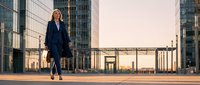 Professional businesswoman walking confidently through modern urban plaza at sunset with glass skyscrapers and contemporary architecture in background.