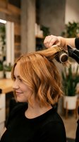 Professional hairstylist using round brush and blow dryer to style blonde woman's bob haircut in modern salon with natural lighting and plants.
