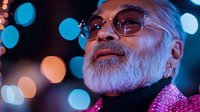Mature Asian man with gray beard and glasses looking up thoughtfully against blurred bokeh lights, wearing pink sweater, modern urban nighttime portrait.
