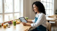 Young Hispanic woman artist working on digital tablet at wooden desk with art supplies and green smoothie in bright studio workspace.