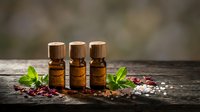 Three amber glass essential oil bottles with wooden caps arranged on rustic wood surface with fresh mint leaves and pink peppercorns for aromatherapy.