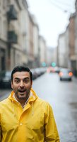 Man wearing bright yellow raincoat on wet urban street during rainy weather. Middle-aged male with dark hair and facial hair, expressing surprise or excitement outdoors in city.