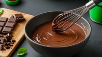 Melted chocolate in ceramic bowl with whisk, dark chocolate pieces and mint leaves on black background. Food preparation for baking and desserts.