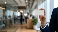 Businessman in dark suit holding blank white business card in modern office lobby with blurred colleagues and glass partitions in background.